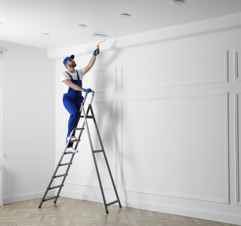 Worker on ladder applying white paint to ceiling in modern interior room