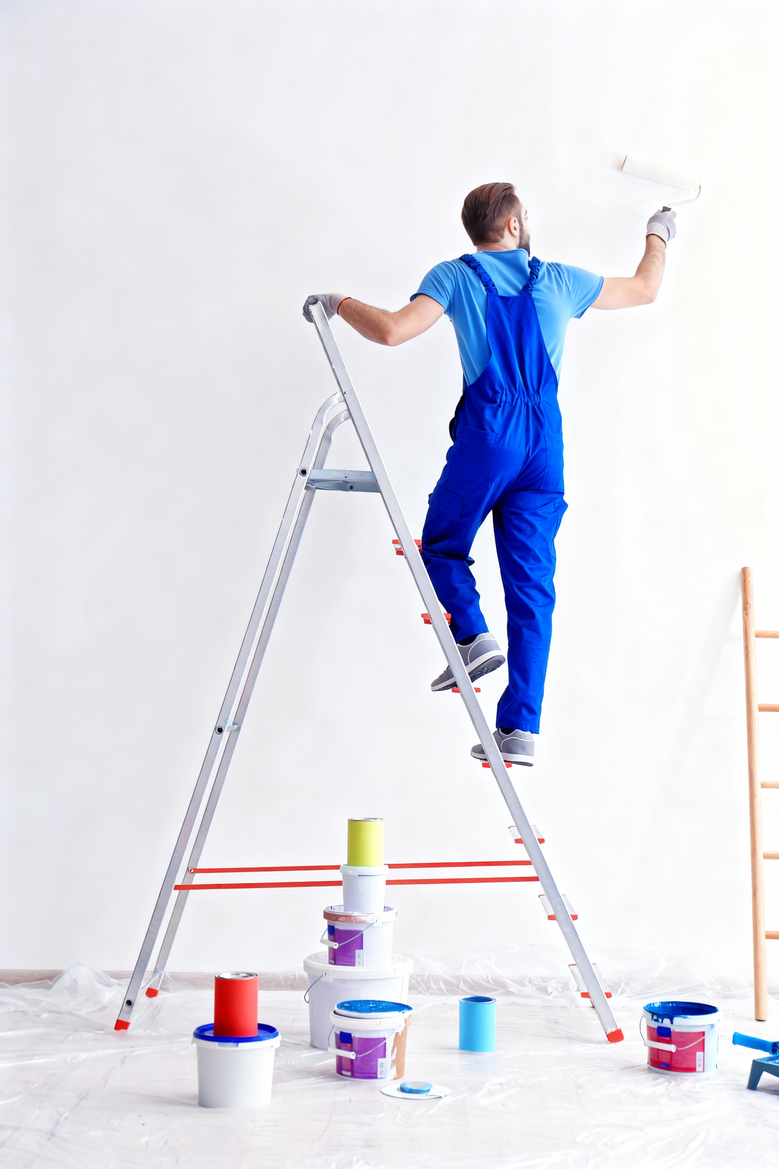 Professional house painter in blue overalls painting a white interior wall with a roller while standing on a ladder, home renovation and painting service