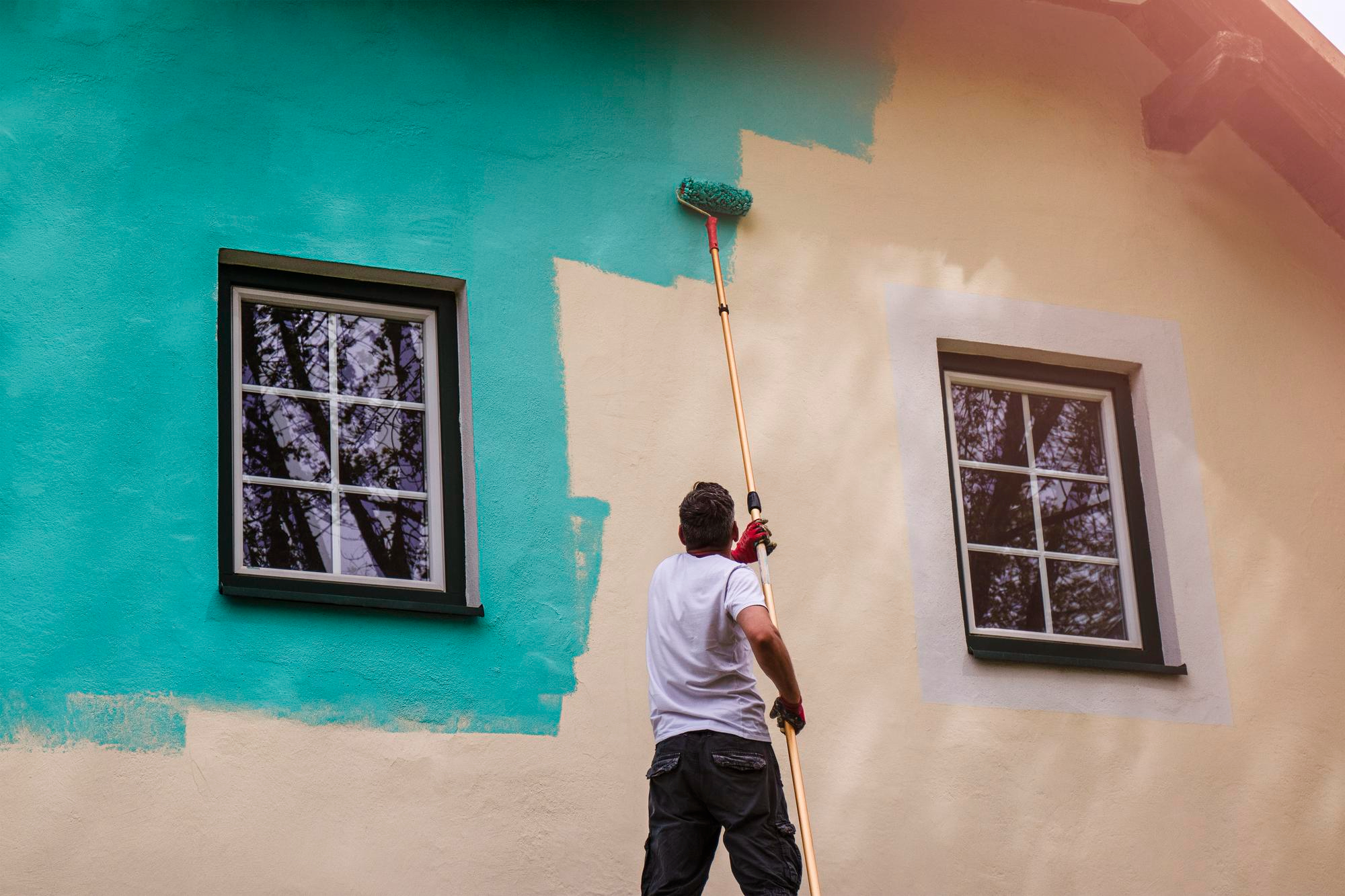 Professional exterior house painter applying teal paint to a home’s stucco wall with a roller